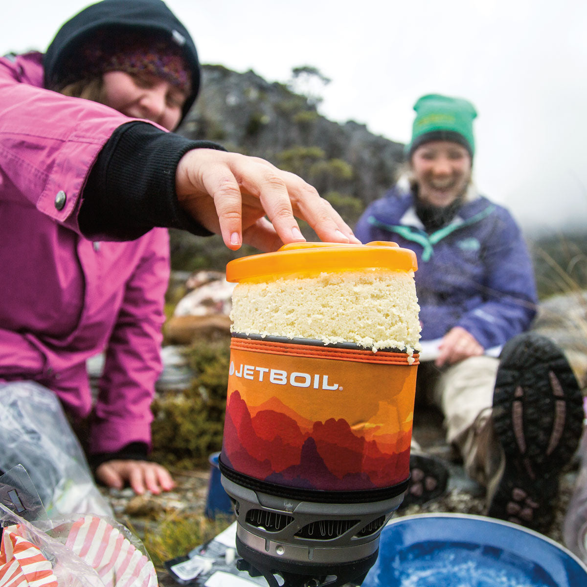 Person holding a JetBoil cooking system outdoors with another person in the background.