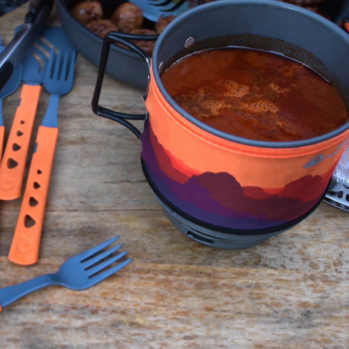 Camping mug with scenic design filled with a beverage, surrounded by utensils on a wooden surface.
