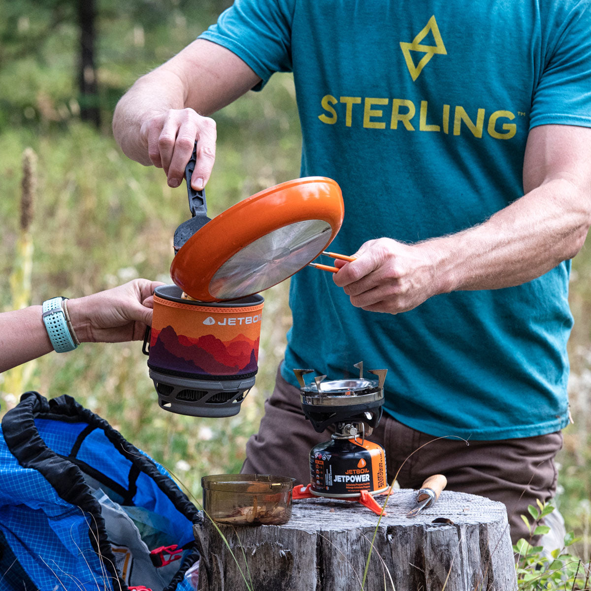 Person cooking outdoors using a Jetboil stove with 'Sterling' branded shirt