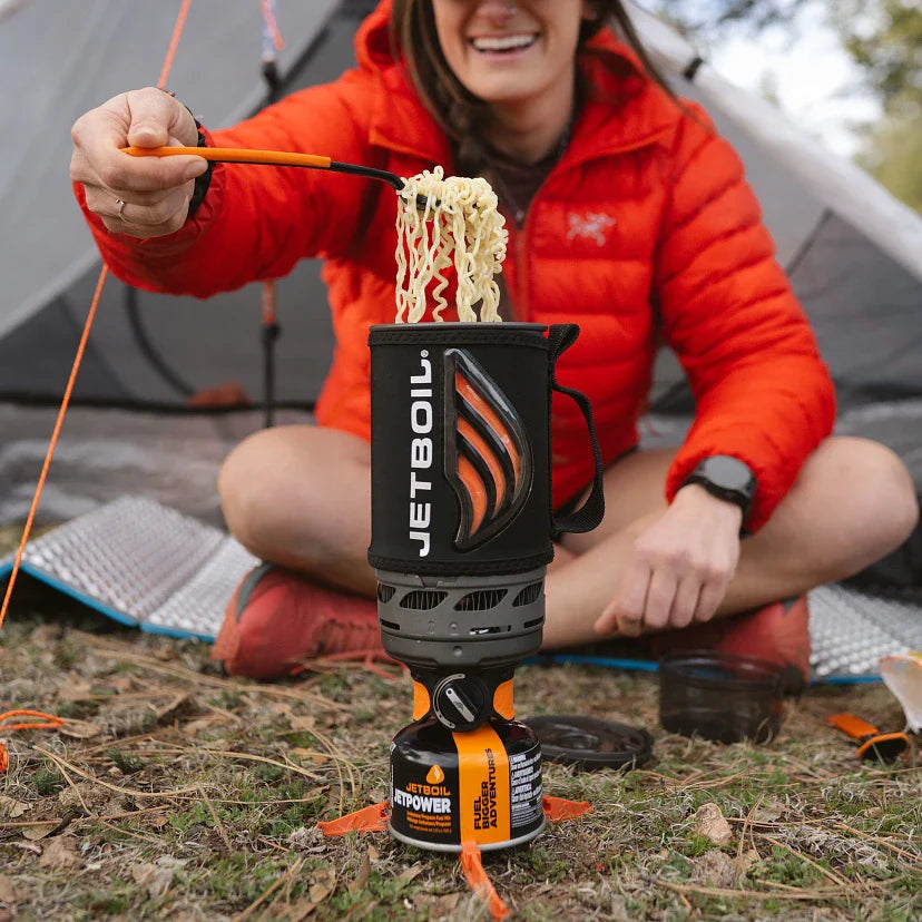 Person cooking noodles using a JetBoil stove outdoors.