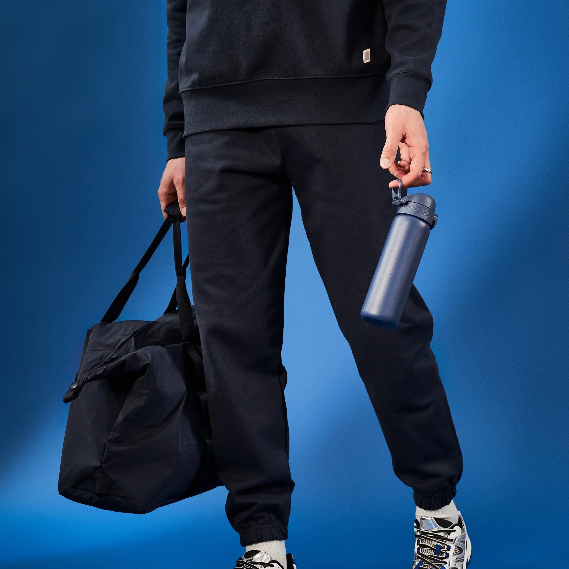 Person wearing black pants holding a black bag and blue water bottle against a blue background