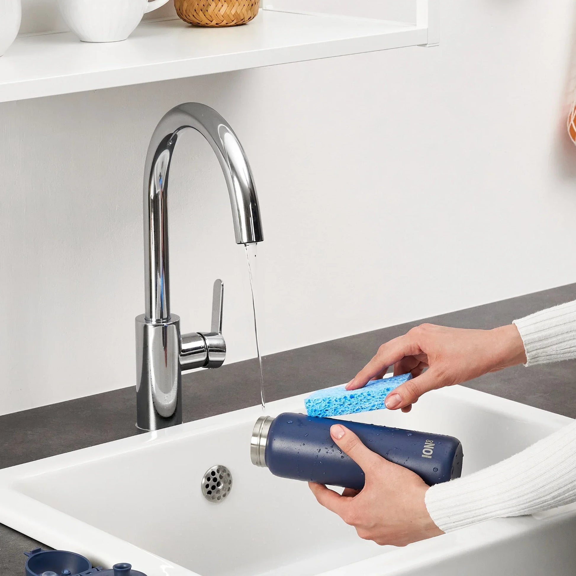 Person cleaning a blue water bottle with a sponge under running water in a kitchen sink.