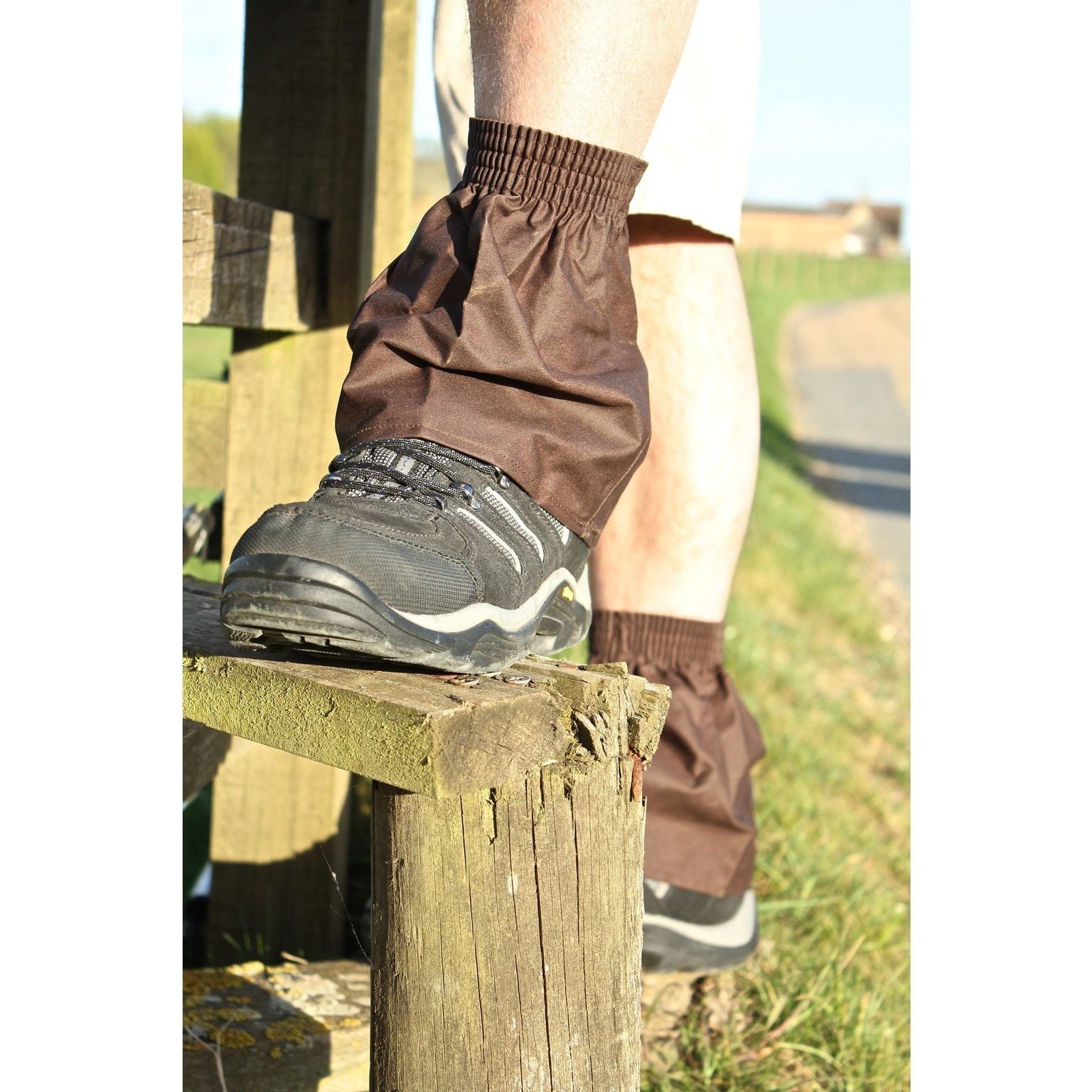 Person wearing brown leg gaiters over black shoes on a wooden post with grass and sky in the background