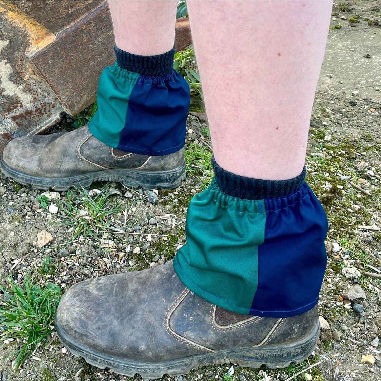 Person wearing green and blue gaiters over work boots on a ground surface.