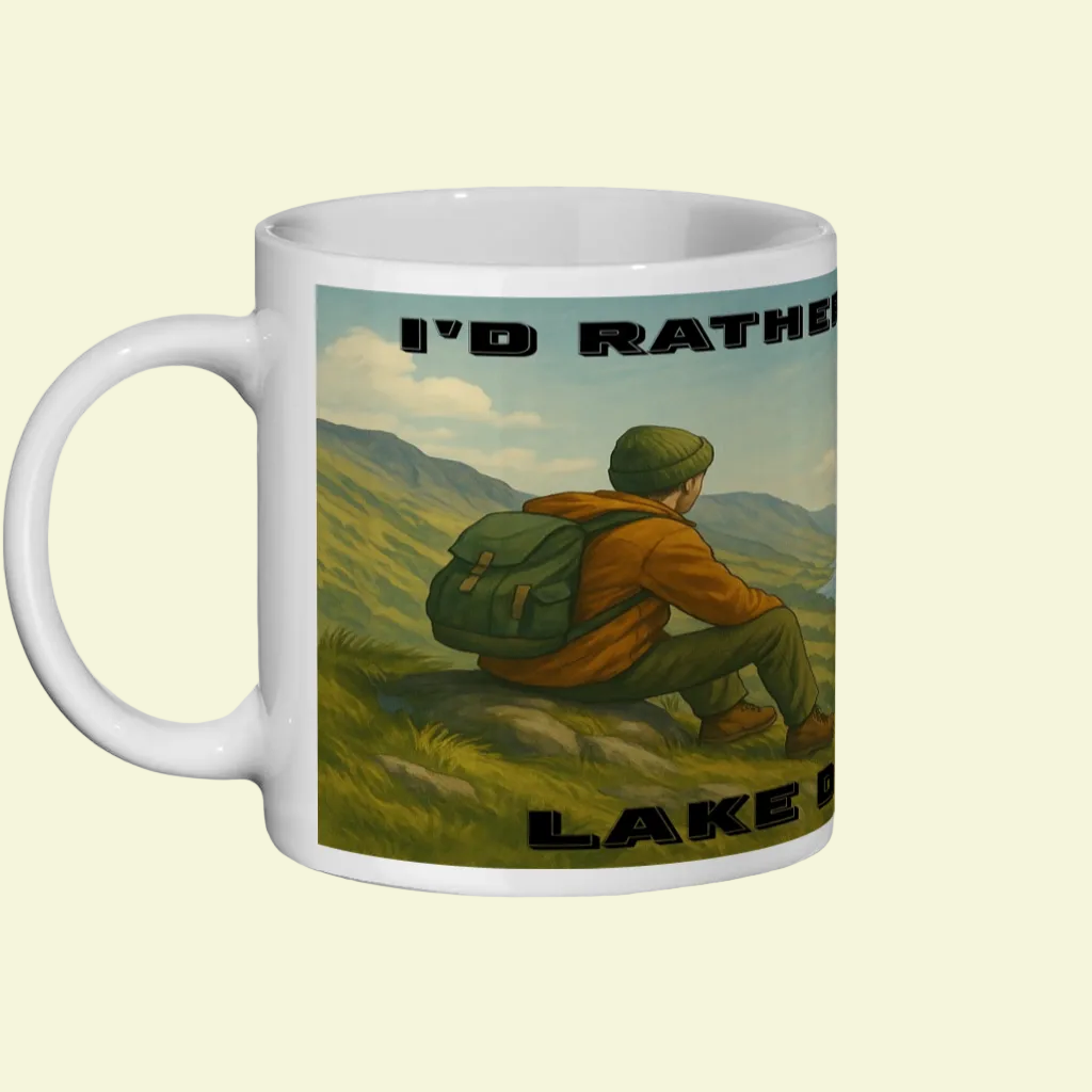 White ceramic mug with illustration of a hiker sitting on a hill overlooking mountains and text "I'd Rather Be in the Lake.