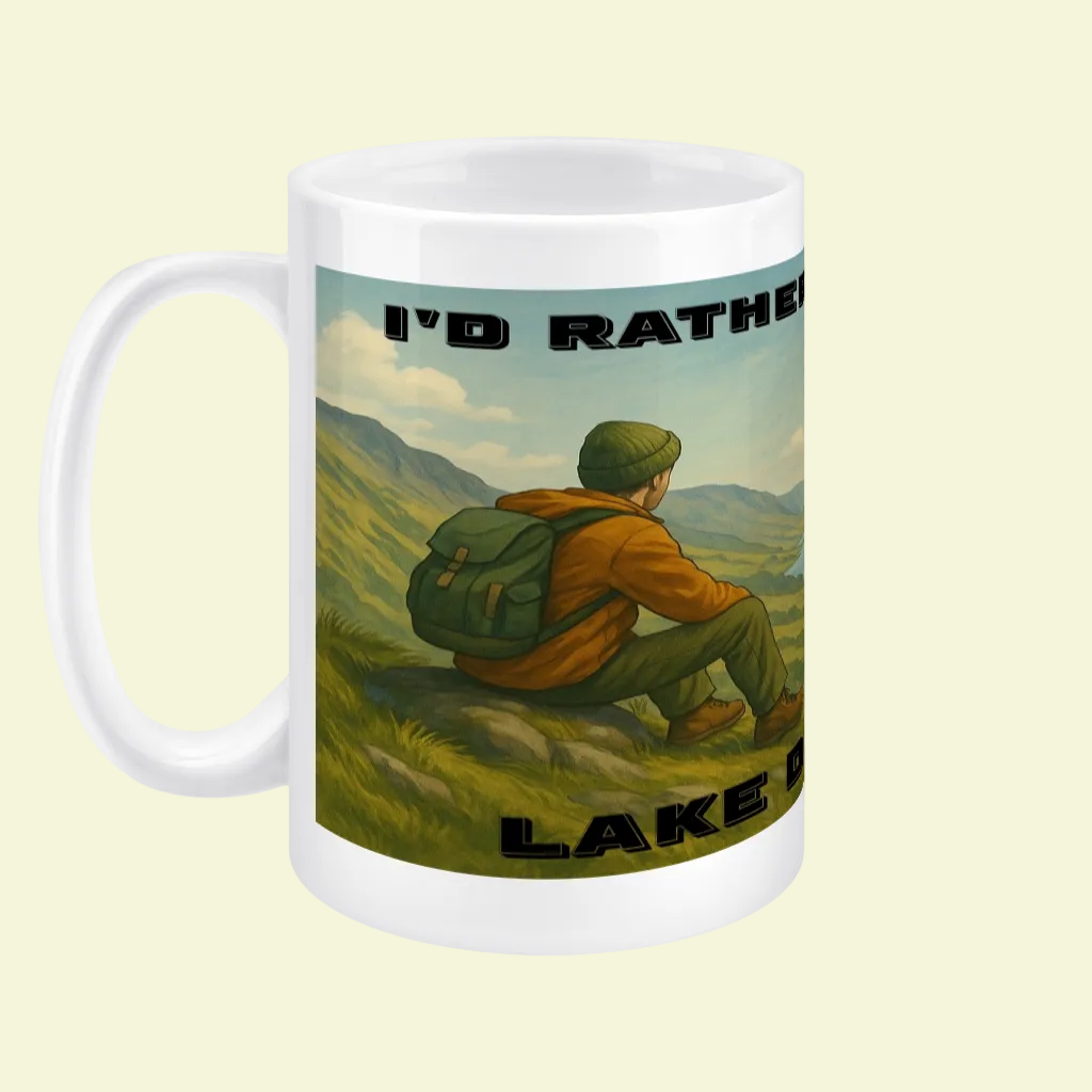 White ceramic mug with a hiker sitting on grass overlooking Lake District hills and black text design.